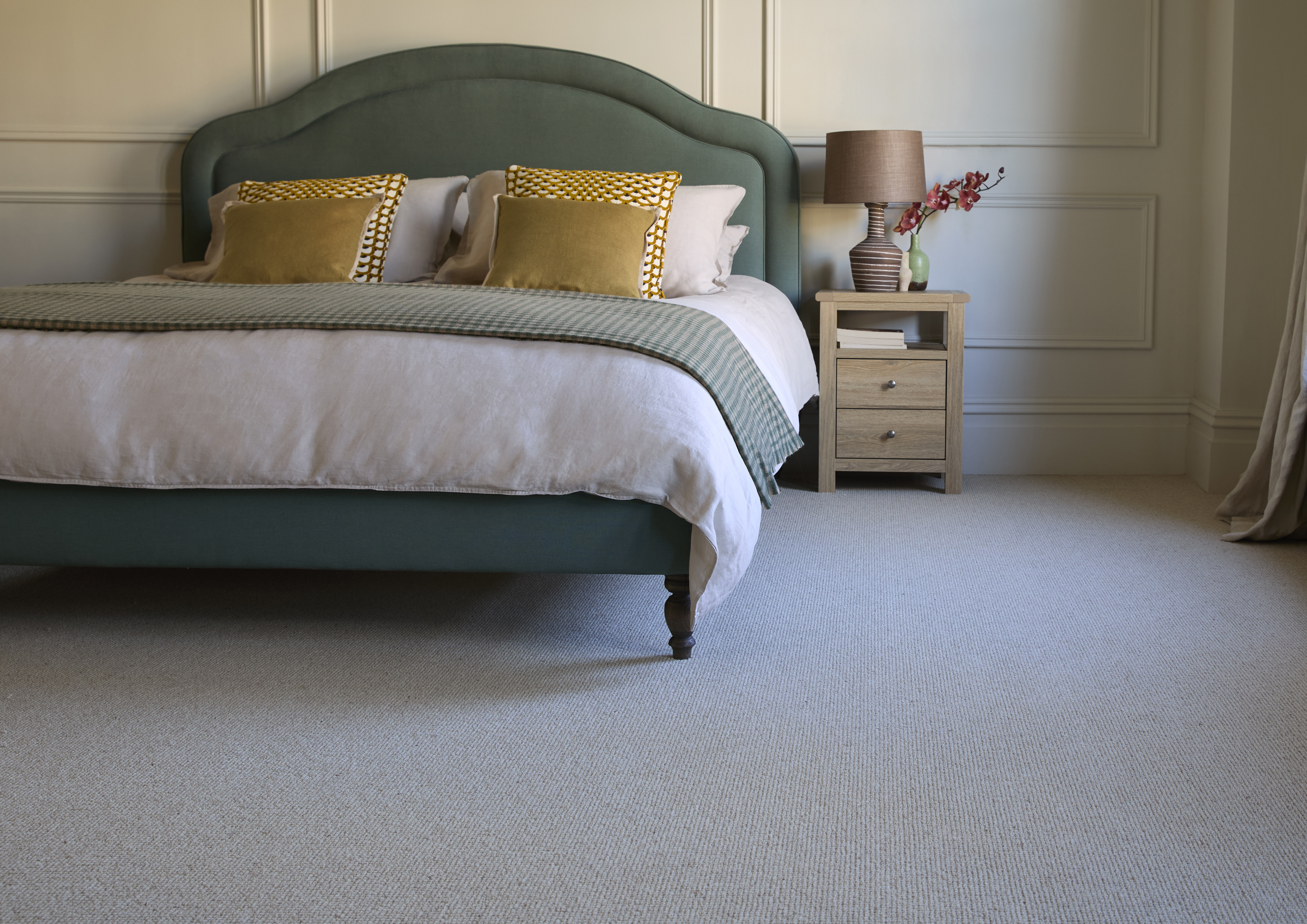 Neutral textured carpet in an elegant master bedroom featuring a sage green upholstered bed, mustard cushions, and light wood bedside table.